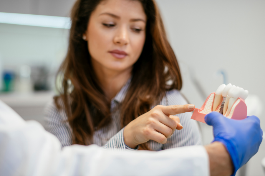 Pacjentka u dentysty wskazująca palcem na model anatomiczny szczęki z implantem zębowym, trzymany przez stomatologa w niebieskiej rękawiczce.