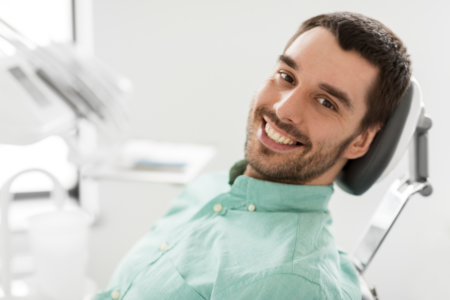 Uśmiechnięty młody mężczyzna siedzi na fotelu dentystycznym w jasnym gabinecie stomatologicznym.