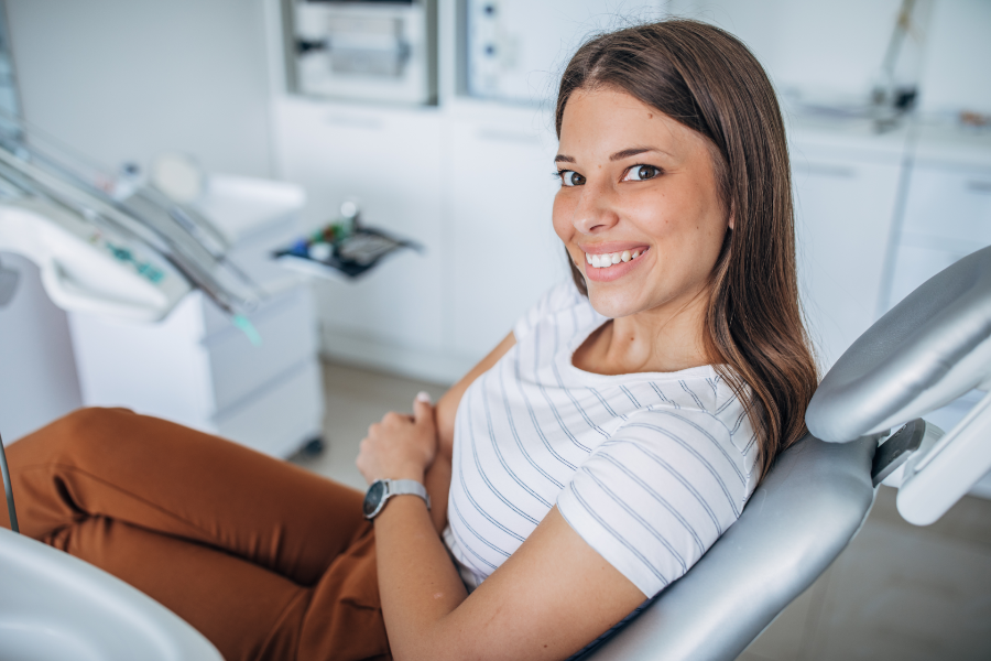 Młoda, brązowowłosa kobieta z białym uśmiechem siedzi na fotelu dentystycznymi patrzy w stronę obiektywu. Ma na sobie białą koszulkę w paski i brązowe spodnie. W tle nowoczesny, jasny gabinet z profesjonalnym sprzętem.
